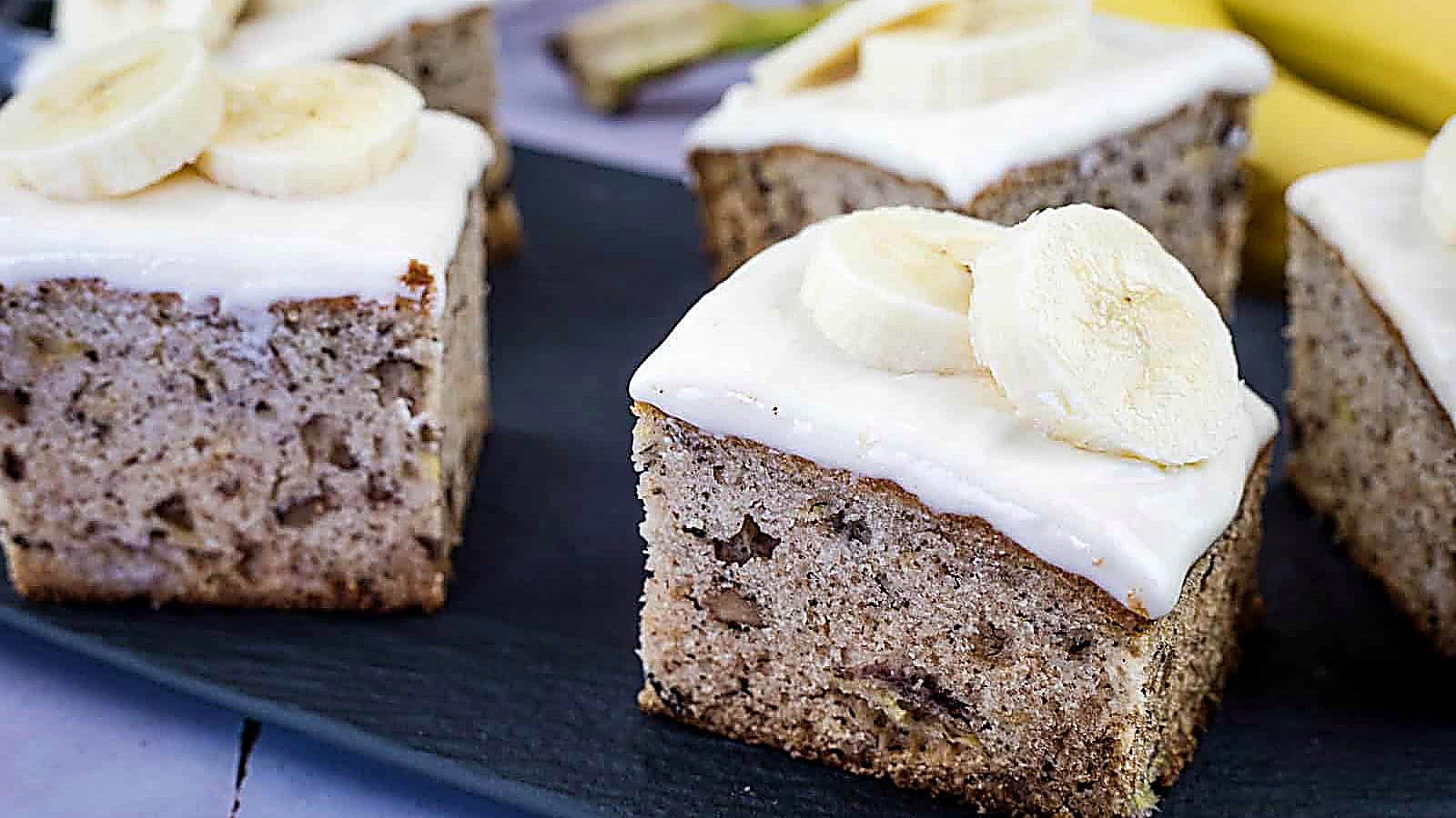 Squares of banana cake topped with white frosting and banana slices are arranged on a dark serving tray, with bananas visible in the background.