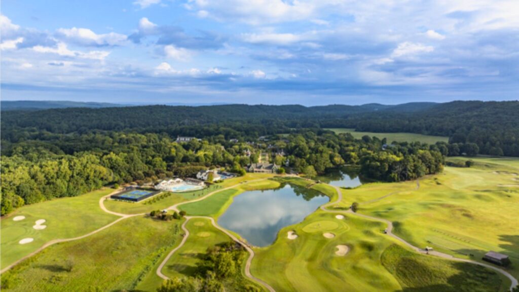 Aerial view of a golf course with sand bunkers, water features, tennis courts, a swimming pool, and surrounding forest under a partly cloudy sky.