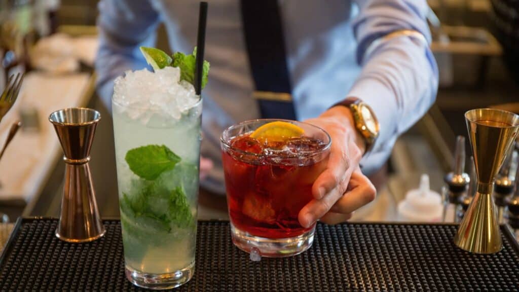 A bartender, mindful of state alcohol laws, serves two cocktails on a bar counter: a tall glass with mint and ice, and a short glass with a dark drink and orange slice.