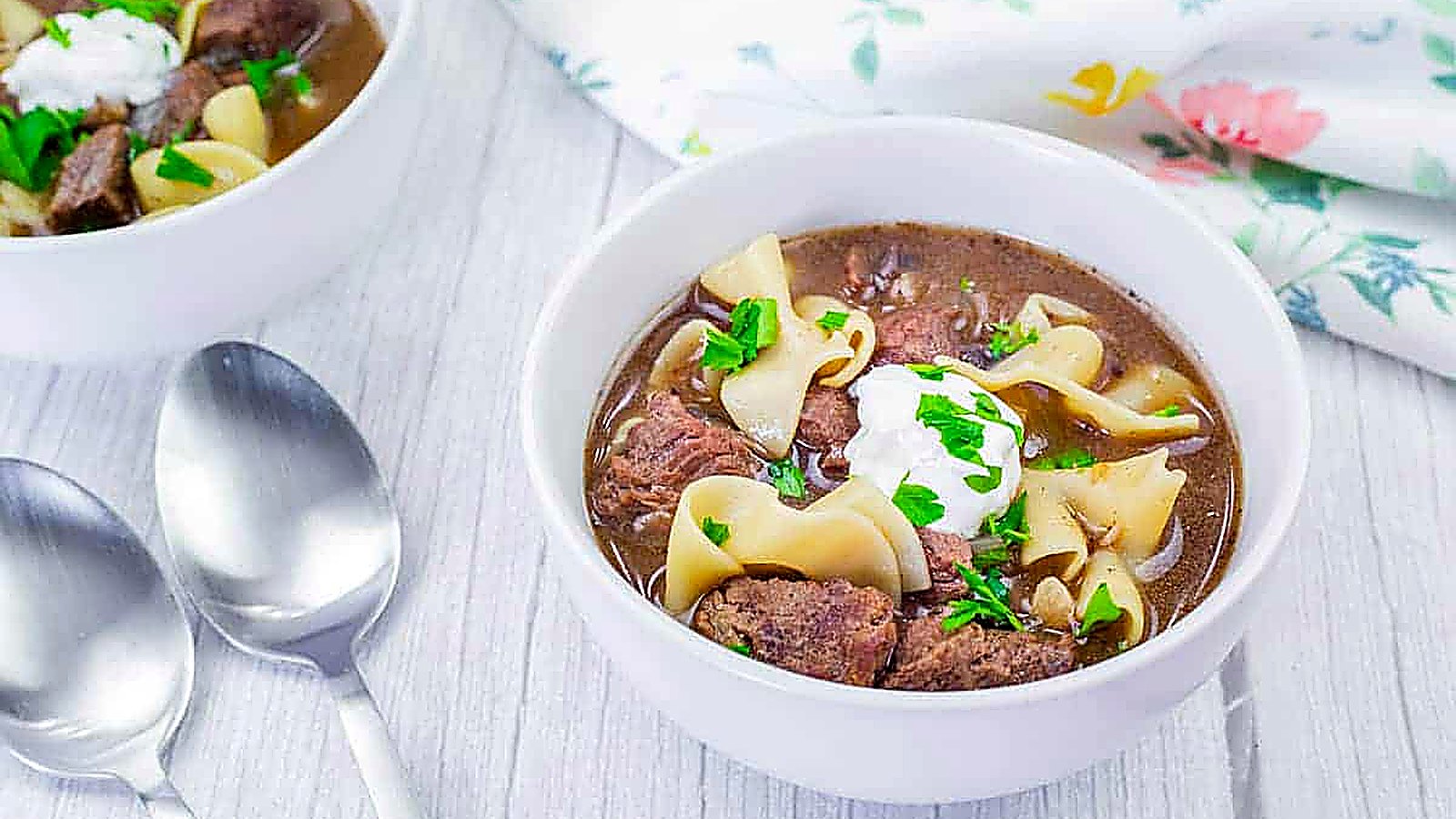 Bowl of beef stroganoff soup with sour cream and parsley garnish on a light wooden table next to two spoons and a floral napkin.