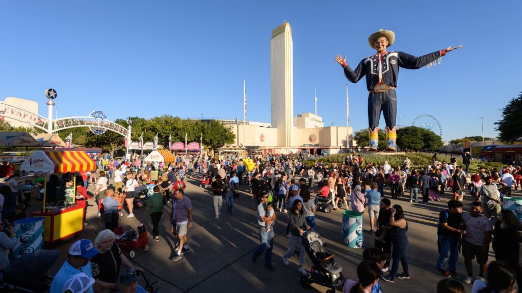 Large crowd at an outdoor fair with food stands, a ferris wheel, and a tall cowboy statue under a clear blue sky, as Texas State Fair finalists are announced amidst the festivities.