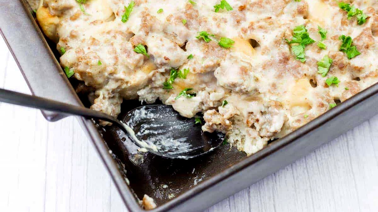 A baking dish of sausage and biscuit casserole topped with creamy gravy and chopped parsley, with a serving spoon partially in the dish.