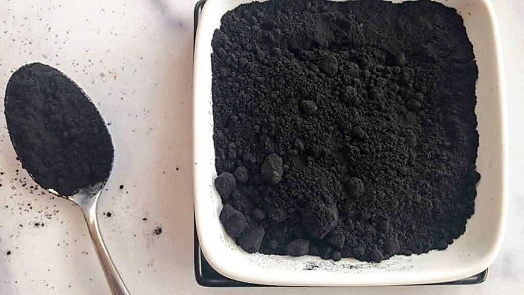 A white dish filled with black powder sits on a table, with a metal spoon holding some of the powder beside it.