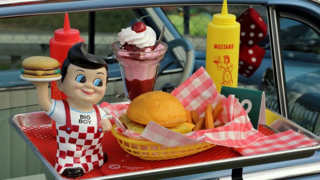 A drive-in food tray holds a Big Boy figurine, burger, fries, ketchup and mustard bottles, and an ice cream sundae set on a car window—a nostalgic scene from regional chains that rarely cross state lines.