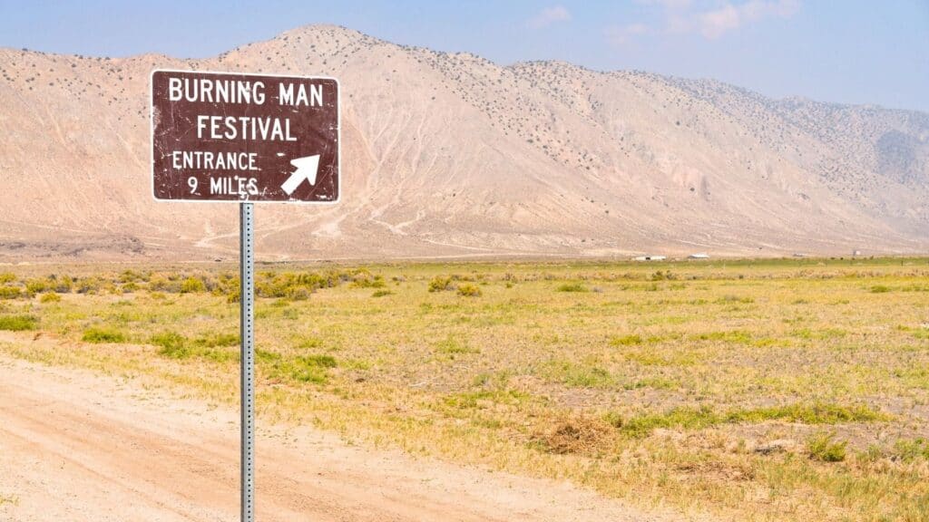 A brown sign by a dirt road in a dry, open landscape reads, &ldquo;Burning Man Festival Entrance 9 Miles,&rdquo; marking the way to Burning Man in Nevada desert, with mountains rising in the background.