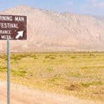 A brown sign by a dirt road in a dry, open landscape reads, “Burning Man Festival Entrance 9 Miles,” marking the way to Burning Man in Nevada desert, with mountains rising in the background.