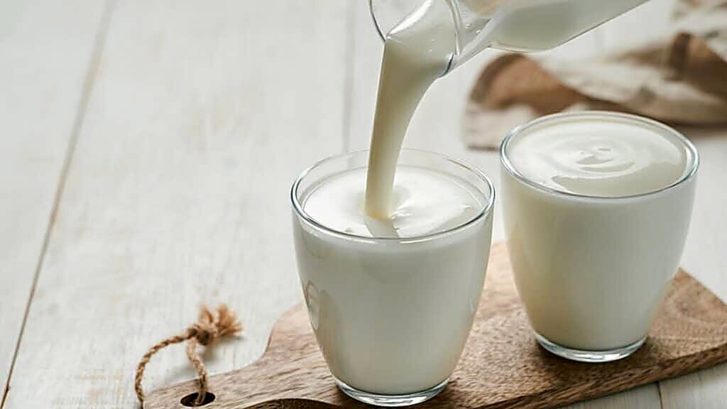 A glass of milk is being poured from a jug next to another full glass of milk, both on a wooden board on a white table.