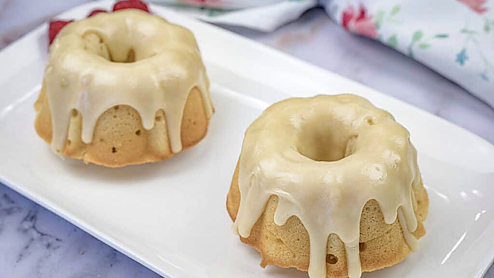 Two small Bundt Grandma's Buttermilk Cake with a smooth white glaze sit on a rectangular white plate.