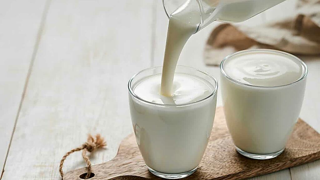 A glass pitcher pours milk into one of two clear glasses placed on a wooden board, with a light-colored surface in the background.