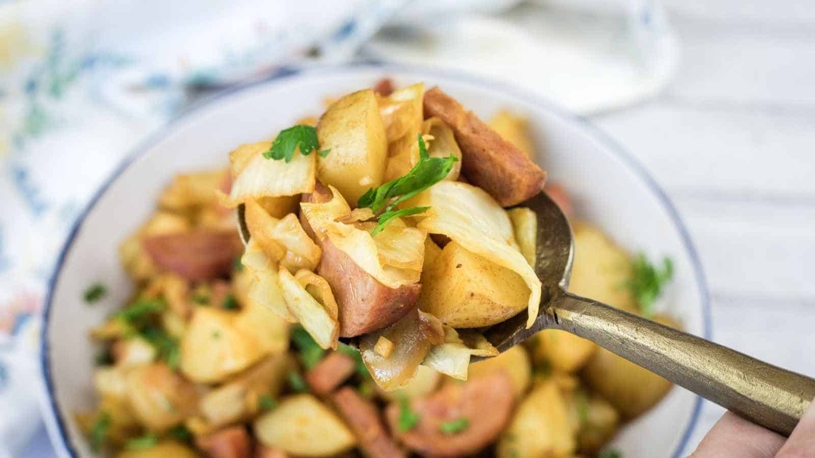 A spoonful of cooked cabbage, potatoes, and sausage held above a bowl filled with the same mixture.