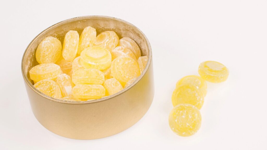 A round container filled with yellow hard candies, reminiscent of candy snacks you can’t buy anymore, with three candies placed outside the container on a white surface.