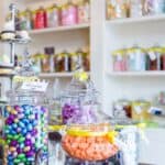 Glass jars filled with colorful candies and chocolates are arranged on a counter in a candy store, including some nostalgic candy snacks you can&rsquo;t buy anymore, with shelves of more jars in the background.
