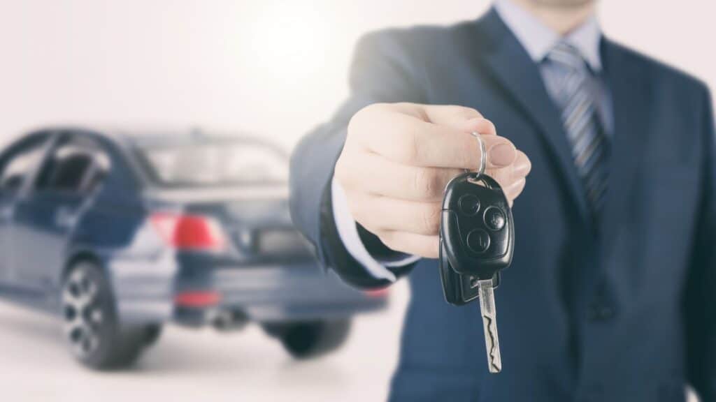 A person in a suit holds out a car key with a sedan in the background, reminding us how unexpected rental car fees catch travelers off guard.