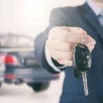 A person in a suit holds out a car key with a sedan in the background, reminding us how unexpected rental car fees catch travelers off guard.