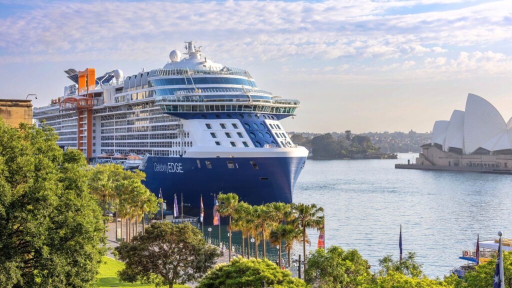 A large Celebrity Cruises ship is docked at a harbor with the Sydney Opera House visible in the background on a partly cloudy day, offering an exciting glimpse into what guests can experience on Celebrity Cruises 2028 and 2029 sailings.