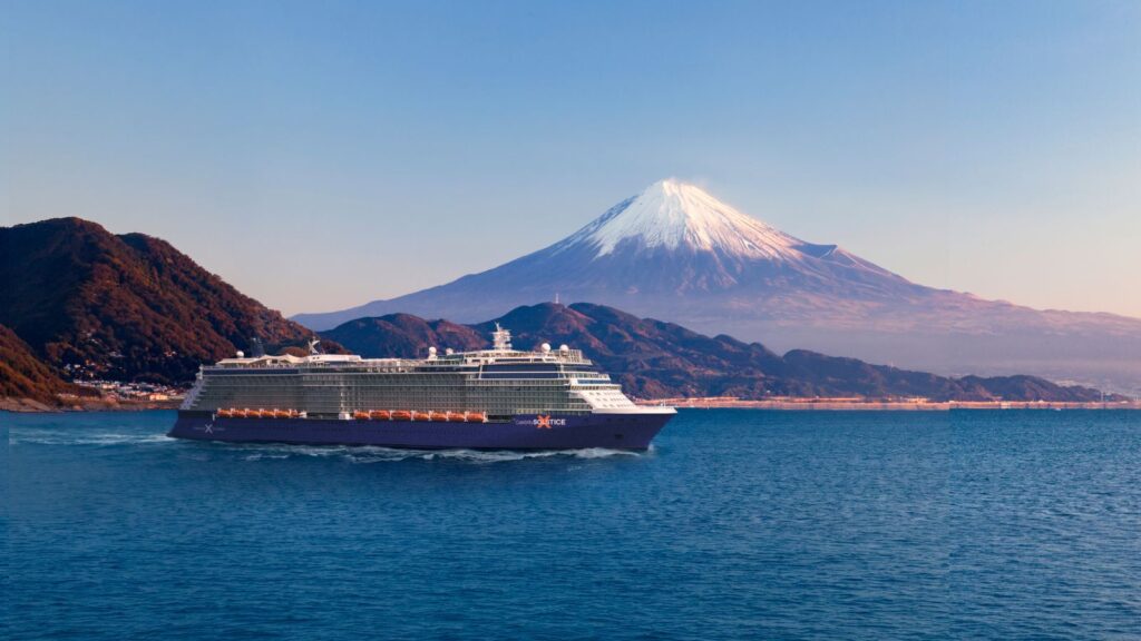 A Celebrity Cruises 2028 or 2029 ship sails on calm blue water with snow-capped Mount Fuji and green hills in the background under a clear sky.