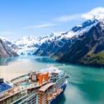 A large Celebrity Cruises ship sails through a turquoise fjord, surrounded by snow-capped mountains and glaciers under a blue sky—an unforgettable scene awaiting travelers on Celebrity Cruises’ 2028 and 2029 itineraries.