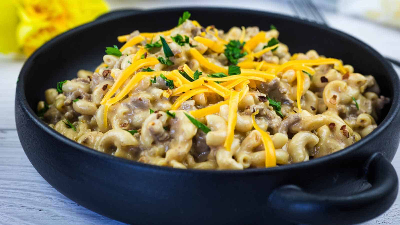 A black bowl filled with cheeseburger hamburger helper topped with shredded cheddar cheese and chopped parsley sits on a white surface.