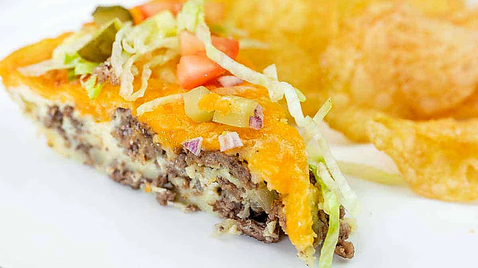 A slice of cheeseburger pie topped with shredded lettuce, diced tomato, onions, and pickles, served with potato chips on a white plate.