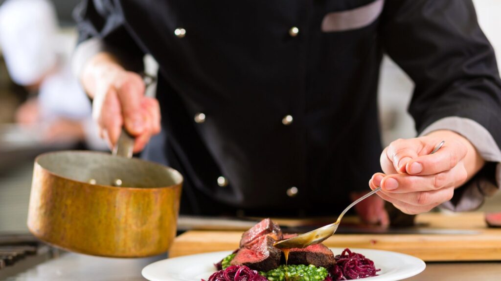 Chef in black uniform plating food, pouring sauce from a saucepan onto a dish with meat, green puree, and red vegetables on a white plate.