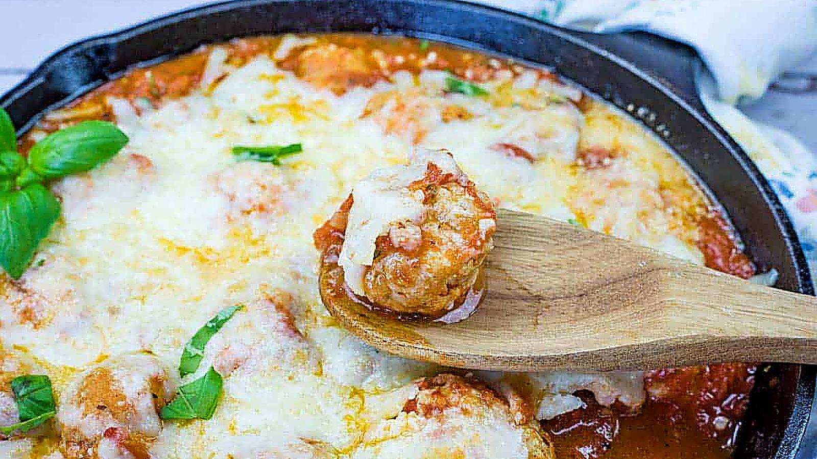 A wooden spoon holds a Chicken Parmesan Meatball over a skillet filled with meatballs.