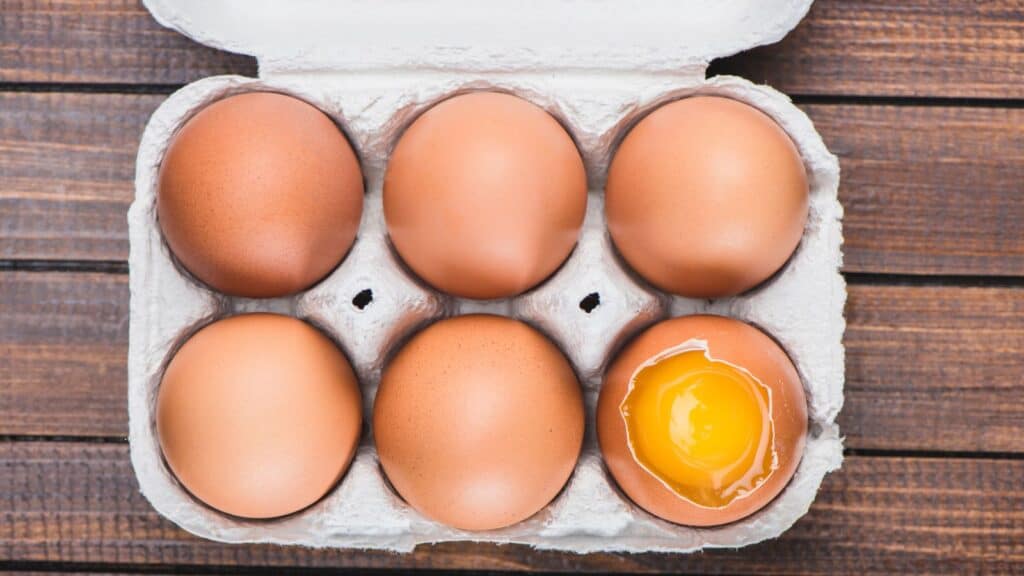 A carton of six brown eggs on a wooden surface, with one egg cracked open to reveal the raw yolk and white, reminds us how important it is to understand what things like a use-by date mean when checking egg freshness.