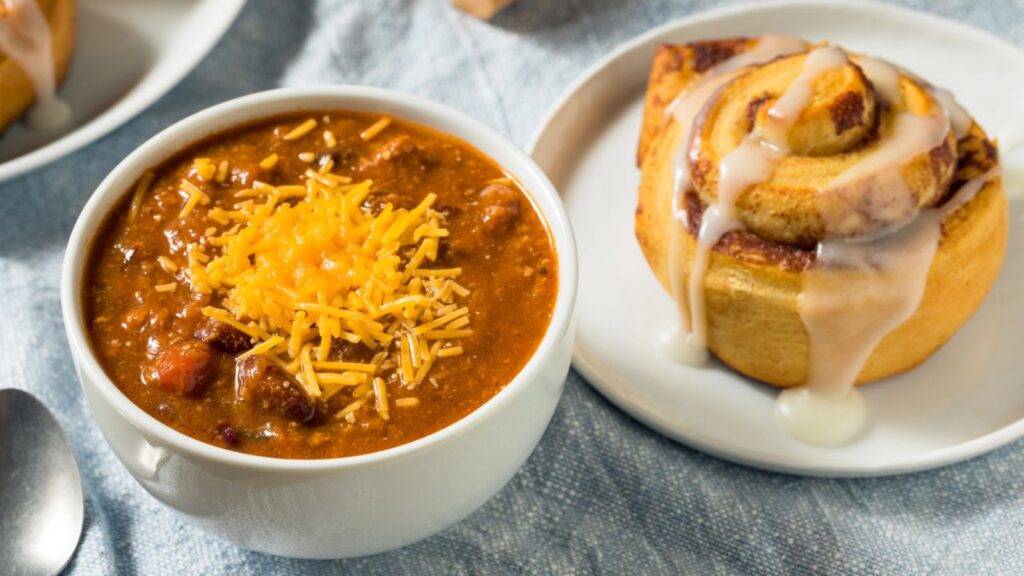 A bowl of chili topped with shredded cheese sits next to an iced cinnamon roll on a white plate, both on a light blue cloth background&mdash;a nostalgic pairing reminiscent of classic 80s and 90s school cafeteria foods.