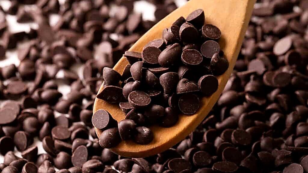 A wooden spoon holds a pile of dark chocolate chips, with more chocolate chips scattered in the background.