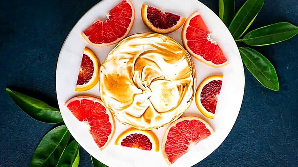 A round meringue-topped dessert sits on a white plate, surrounded by grapefruit wedges. Green leaves are placed around the plate on a dark surface.