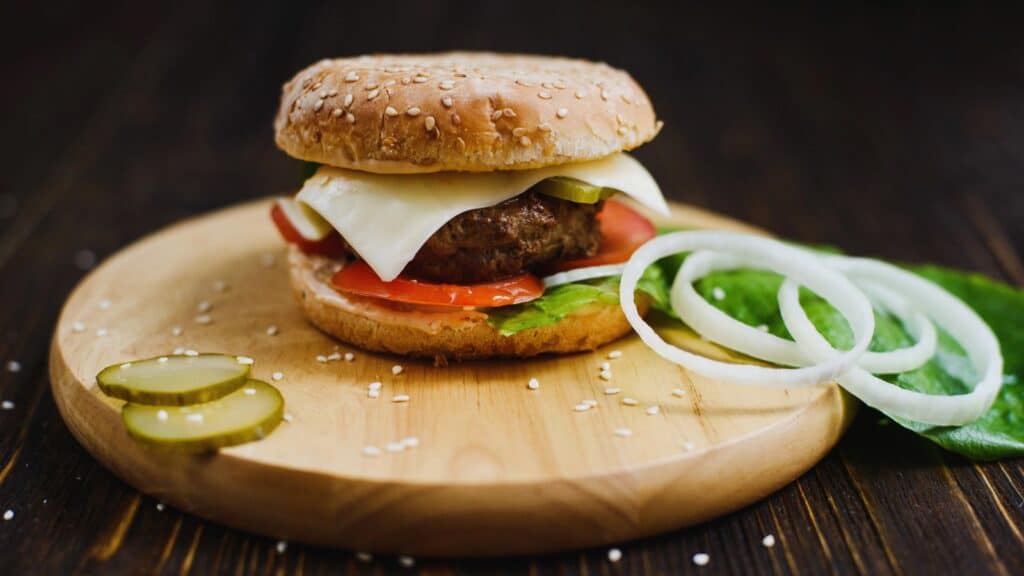 A cheeseburger with lettuce, tomato, onions, pickles, and cheese on a sesame bun sits on a wooden board, accompanied by extra onion rings and pickle slices on the side&mdash;a delicious example of American foods with foreign roots.