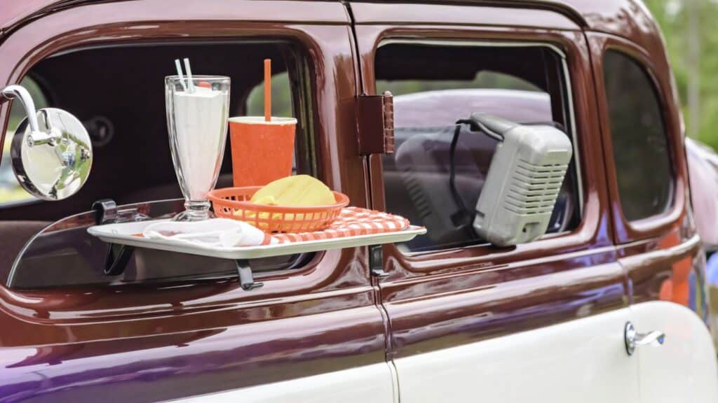 A classic car with a tray on the window holding a milkshake, a drink cup, and a basket of food sits ready to serve up drive-in snack-bar oddities, complete with a retro drive-in speaker attached to the window.
