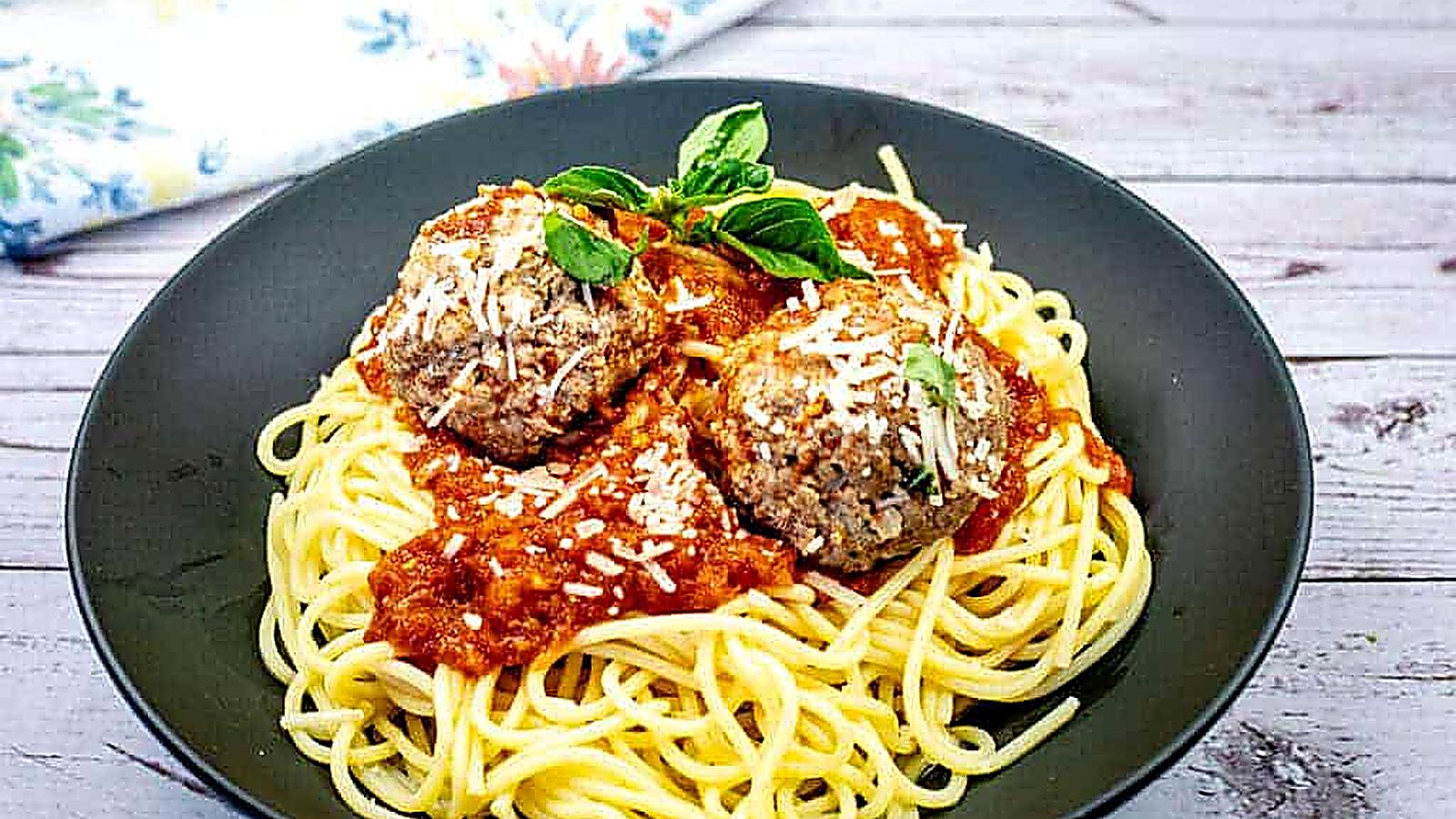 A plate of spaghetti topped with two meatballs, marinara sauce, grated cheese, and basil garnish.