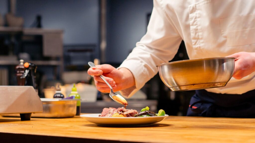 A chef in a white uniform uses a spoon to add sauce to a plated dish on a wooden counter in a professional kitchen.