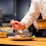 A chef in a white uniform uses a spoon to add sauce to a plated dish on a wooden counter in a professional kitchen.