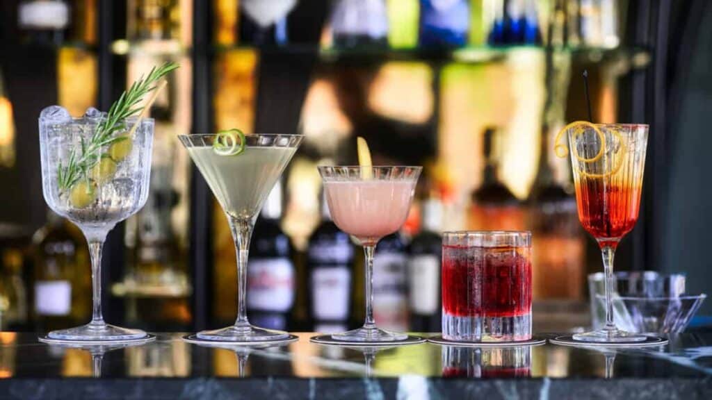 Five assorted cocktails in elegant glasses on a bar counter with a blurred background of liquor bottles.