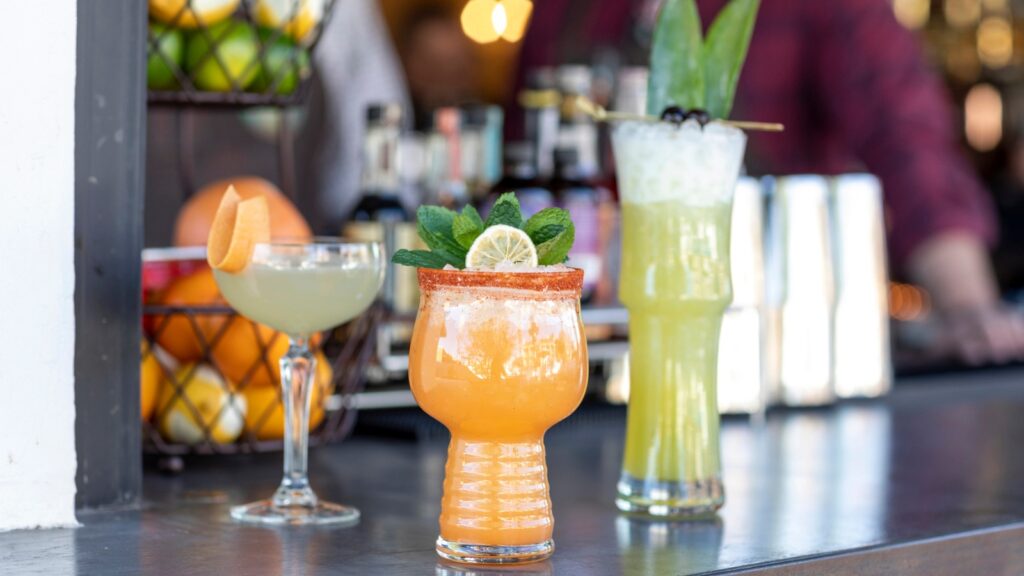Three colorful cocktails on a bar counter with fruit and bottles in the background; one orange drink garnished with mint and lime, one green drink with leaves, and one pale drink with an orange slice.