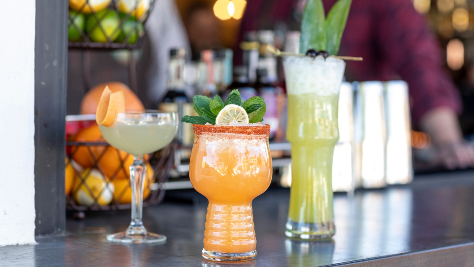 Three colorful cocktails on a bar counter with fruit and bottles in the background; one orange drink garnished with mint and lime, one green drink with leaves, and one pale drink with an orange slice.