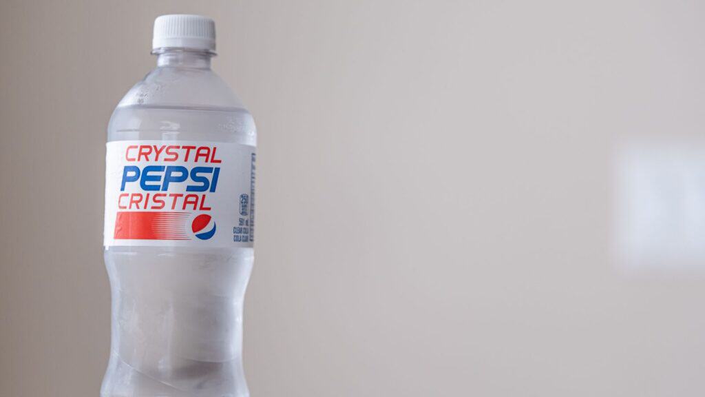 A clear plastic bottle of Crystal Pepsi with a white cap and a label displaying the brand name in red and blue text, against a plain beige background&mdash;a memorable example among sodas that had their moment.