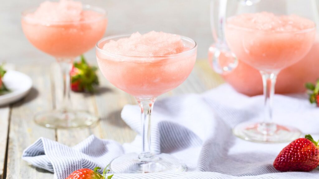 Three coupe glasses filled with pink frozen drinks are on a table with a white cloth and scattered strawberries.