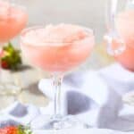 Three coupe glasses filled with pink frozen drinks are on a table with a white cloth and scattered strawberries.