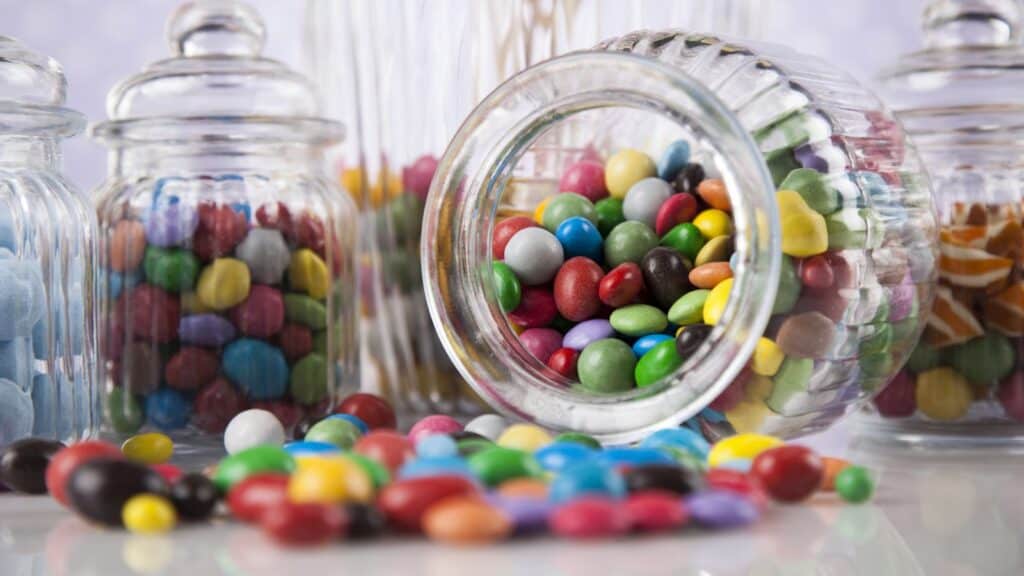 A glass jar tipped over, spilling colorful candy-coated chocolates onto a surface, with other jars of assorted candies in the background.