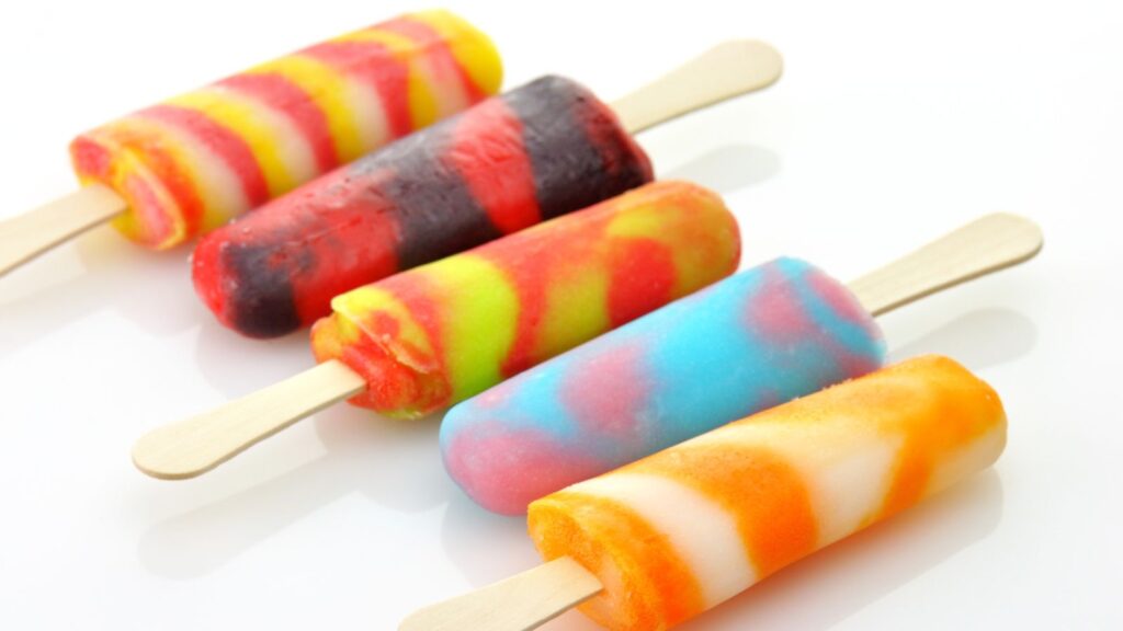 Five colorful swirled popsicles, reminiscent of classic ice cream truck treats, with wooden sticks are arranged diagonally on a white surface.