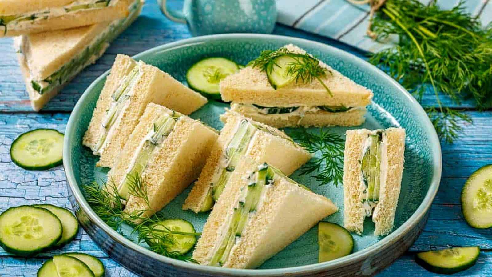 A plate of triangular cucumber sandwiches on white bread, garnished with fresh dill and cucumber slices, set on a blue wooden table.