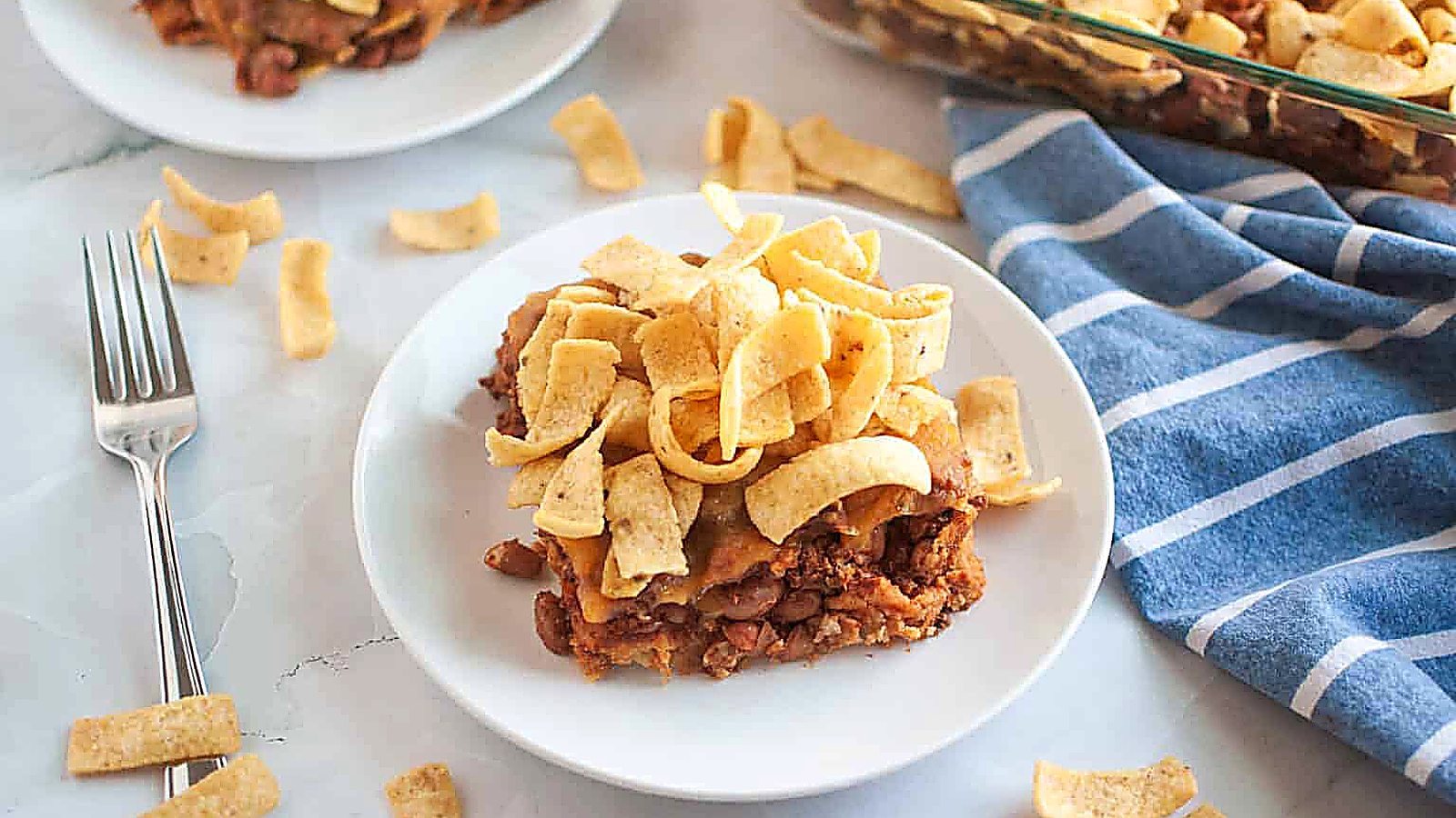 A white plate with a serving of Frito pie, topped with corn chips, is shown on a light surface.