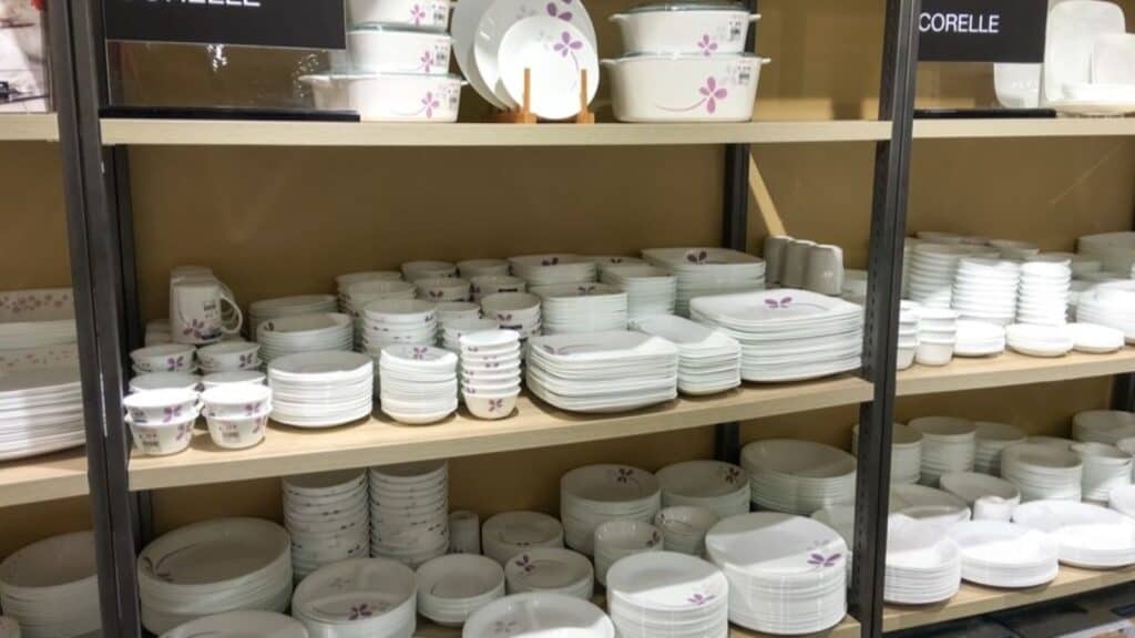 Shelves displaying stacks of white ceramic plates, bowls, and cups with purple floral patterns&mdash;classic kitchen gear Americans remember&mdash;are neatly organized in a store.