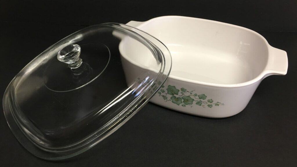 A white ceramic casserole dish with green floral design&mdash;a piece of kitchen gear Americans remember&mdash;stands with its clear glass lid placed beside it, set against a black background.