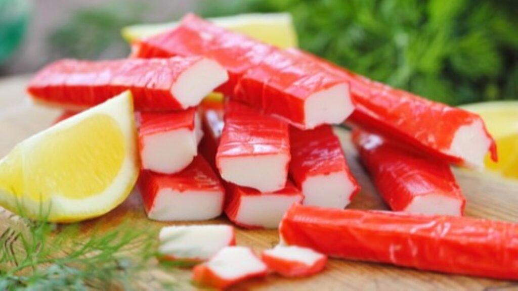 Several pieces of imitation crab sticks&mdash;one of those classic foods not what name suggests&mdash;are stacked on a wooden surface, with lemon wedges and fresh herbs in the background.