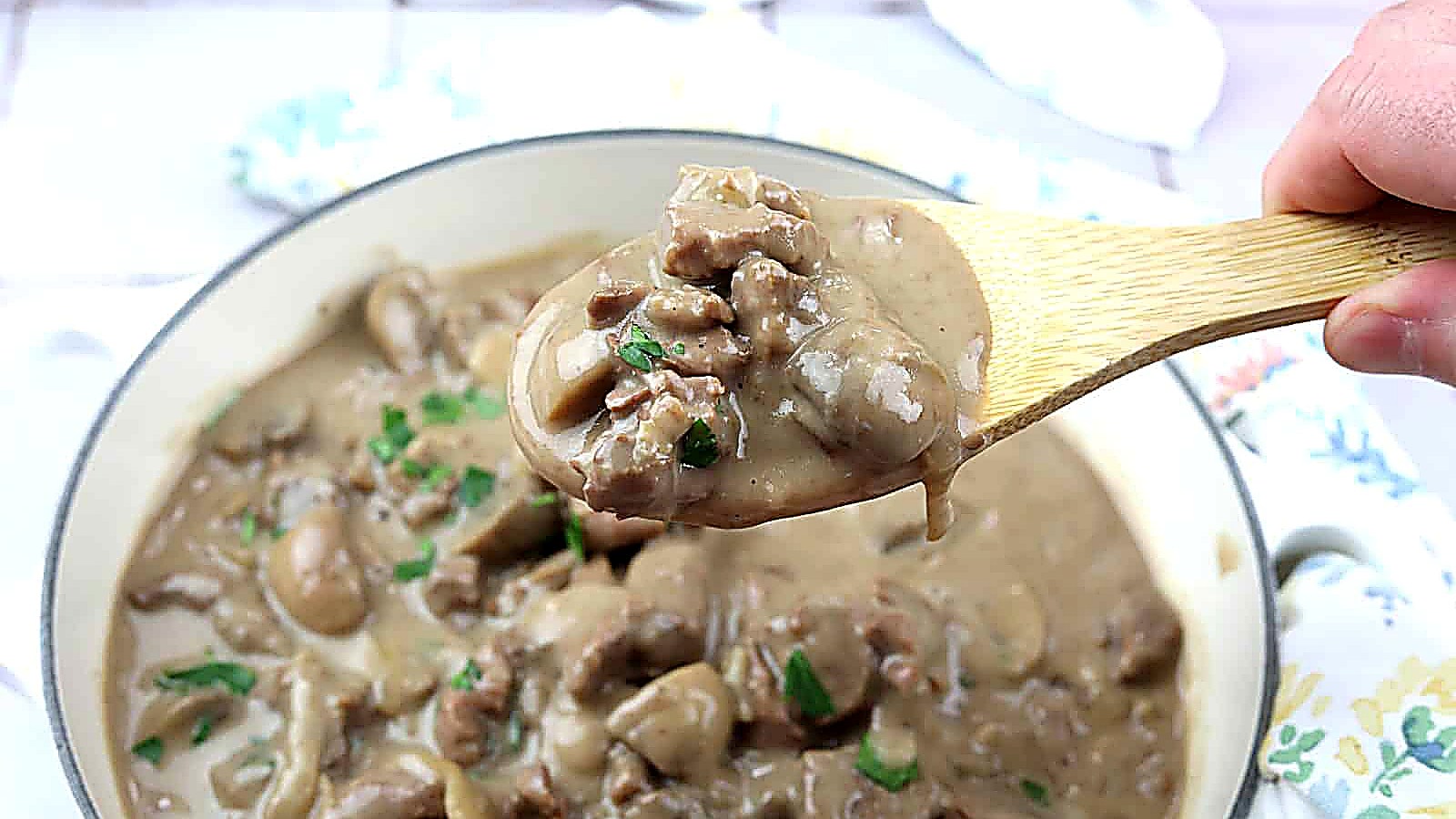 A wooden spoon holds a serving of beef stroganoff with mushrooms and creamy sauce above a pot filled with the dish.