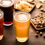 Three glasses of beer on a wooden table, surrounded by dishes of assorted snacks including potato chips, crackers, and pistachios.