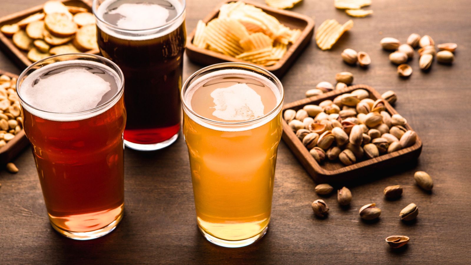 Three glasses of beer on a wooden table, surrounded by dishes of assorted snacks including potato chips, crackers, and pistachios.